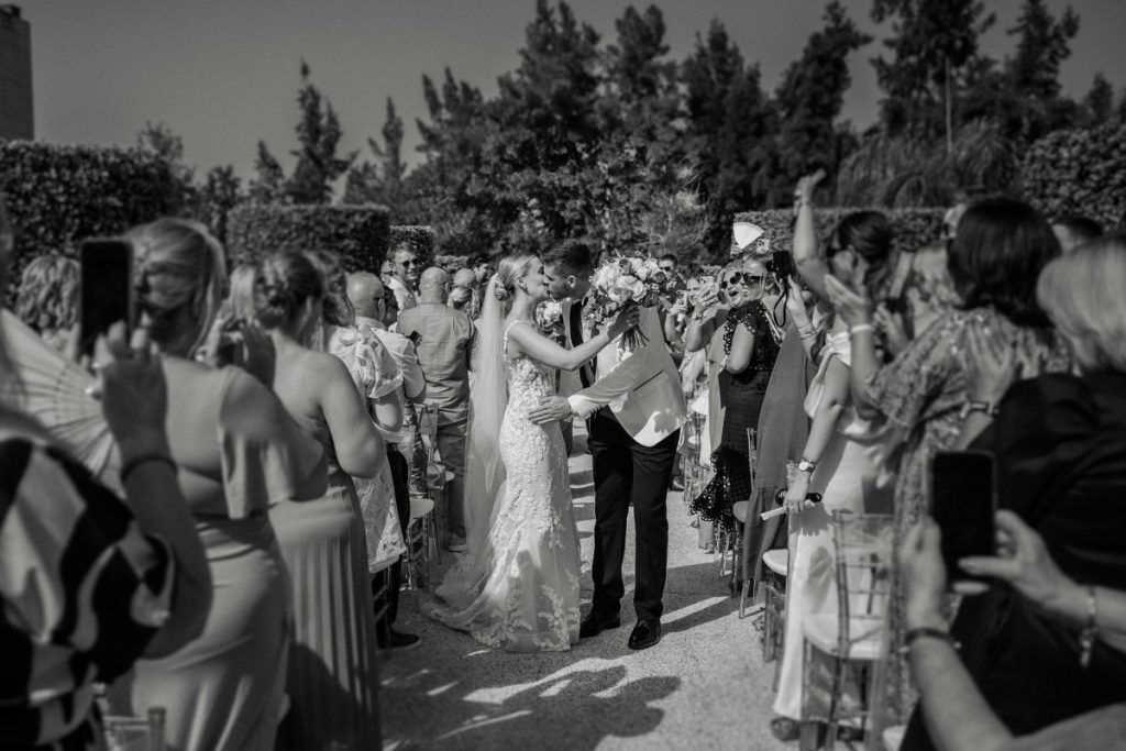 Black-and-white photo of an international couple kissing down the aisle at an outdoor ceremony in La Finca Resort Spain, guests applauding — destination wedding photographer Alicante Costa Blanca.