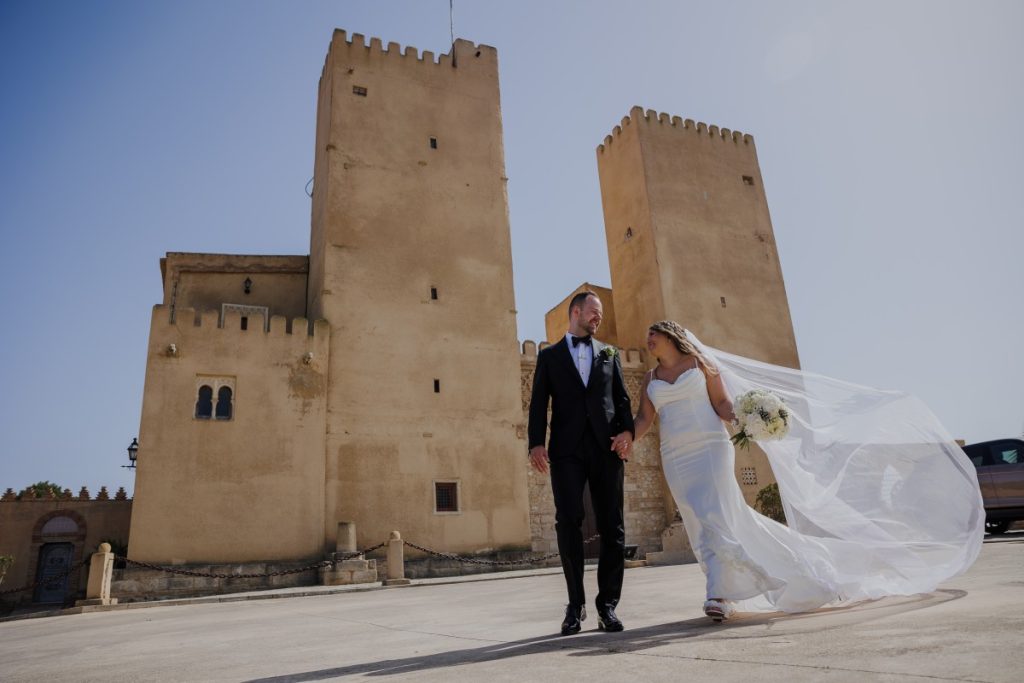 Alicante wedding photographer reviews:Norwegian couple just married in Spain, walking hand in hand with a flowing veil in front of a historic Alingui Castle — destination wedding photographer Alicante Costa Blanca.